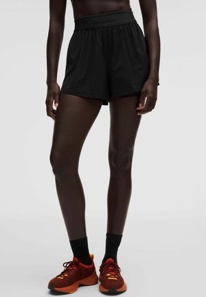 Persoon met zwarte sportshorts, zwarte sokken en rood-oranje hardloopschoenen, staand tegen een effen lichte achtergrond.
