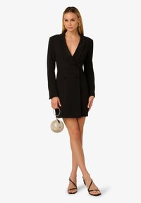 Czarna, dwurzędowa sukienka w marynarskim stylu z głębokim dekoltem w kształcie litery V, ozdobnym kołnierzem, wyraźnie zarysowanymi ramionami oraz dopasowanym krojem, w połączeniu z okrągłą torbą.
