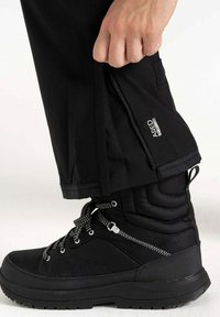 Schwarze, wasserdichte Hose mit seitlichem Reißverschluss, Slim-Fit-Schnitt und elastischen Bündchen, kombiniert mit einem schwarzen gepolsterten Stiefel mit strukturierten Akzenten.