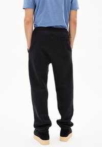 Pantalon de survêtement noir avec une taille élastique et des chevilles resserrées. Comprend une poche arrière et est porté avec des baskets noires à semelles beiges.