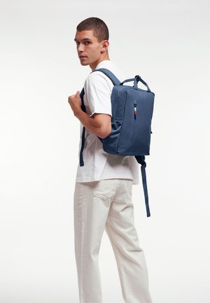Sac à dos bleu marine de forme rectangulaire, en tissu lisse, avec une poche zippée à l'avant et des bretelles réglables. Présente un petit accent multicolore.