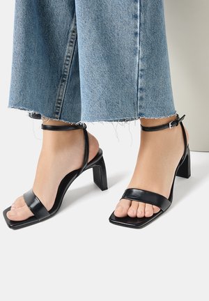 Pies que llevan sandalias de tacón alto negras con correas en el tobillo y correas delanteras anchas, acompañadas de unos jeans de mezclilla azul con dobladillo deshilachado.