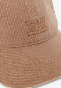 Svetlo rjav klobuk z teksturiranim žametnim materialom. Vezenina logotipa z dvema vodoravnima črtama blizu roba.