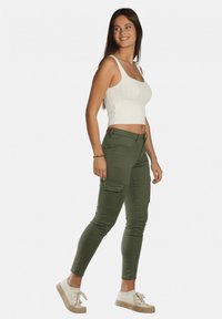 Pantalon cargo vert avec poches latérales et coupe ajustée, associé à un haut court en tricot blanc. Le modèle porte des chaussures beige à enfiler.