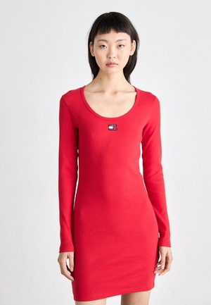 Red long-sleeve fitted dress made of ribbed fabric, featuring a scoop neckline and a small branded tag on the front. Smooth texture.