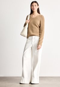 Tan strikket genser med V-hals, kombinert med hvite vidbente bukser. Modellen holder en kremfarget skulderveske og står foran en nøytral bakgrunn.