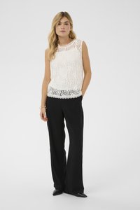 Weißes ärmelloses Spitzenoberteil mit geschwungenem Ausschnitt, über schwarzen, weit geschnittenen Hosen getragen. Strukturierter Stoff mit floralen Akzentmustern.