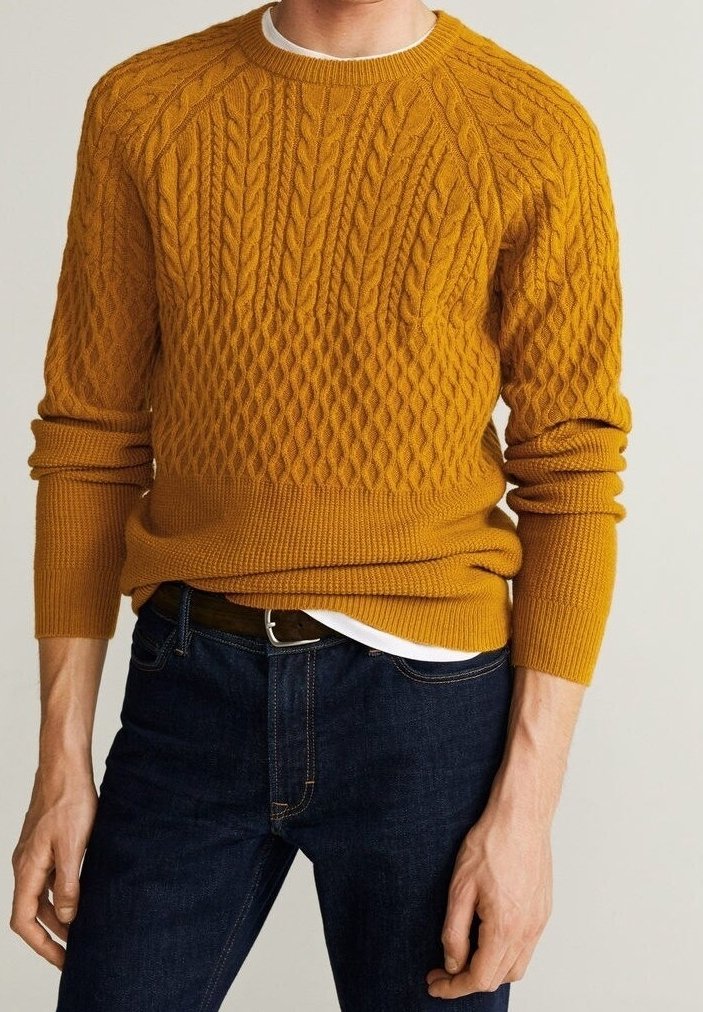Homme portant un pull jaune moutarde à torsades par-dessus une chemise blanche et un jean bleu foncé, debout devant un fond uni.