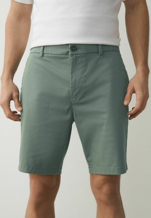 Mann trägt grüne maßgeschneiderte Shorts und ein weißes T-Shirt, steht mit entspannt an den Seiten hängenden Händen vor einem einfarbigen Hintergrund.