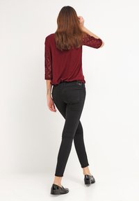 Bordeaux topp med spetsärmar, kombinerad med figurnära svarta jeans och svarta flats, sedd bakifrån mot en neutral bakgrund.