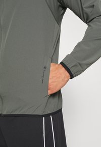 Chaqueta deportiva gris con cierre de cremallera, paneles texturizados y un logo en el lado izquierdo. Cuenta con puños elásticos y un bolsillo lateral.