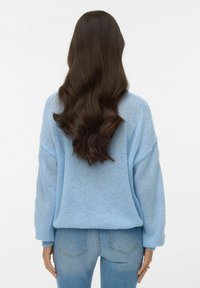 Svetlo modra pletena majica z ohlapnim krojem, spuščene ramena in rebrastimi ovratniki, kombinirana z oprijetimi modrimi kavbojkami. Priložnostna tekstura in slog.