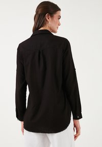 Black long-sleeve shirt featuring a collar, buttoned cuffs, pleat detail at the back, and a curved hem. Made of lightweight fabric.