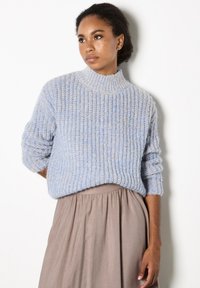 Hellblauer gestrickter Pullover mit hohem Kragen und gerippter Textur, kombiniert mit einem taupefarbenen, fließenden Rock. Der Pullover hat lockere, aufgerollte Ärmel.