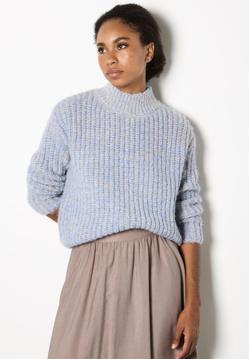 Hellblauer gestrickter Pullover mit hohem Kragen und gerippter Textur, kombiniert mit einem taupefarbenen, fließenden Rock. Der Pullover hat lockere, aufgerollte Ärmel.