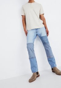 Beige randig henley-skjorta med korta ärmar, i kombination med patchwork jeans i olika blå nyanser, och bruna ankelskor.
