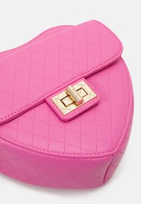 Kabelka ve tvaru srdce, růžová, prošívaná, s lesklým povrchem, vybavená zlatým zapínáním s pyramidovými hroty a hladkým povrchem.