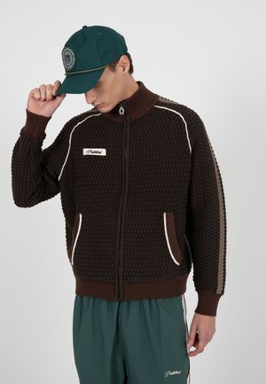 Junger Mann, der eine dunkelgrüne Mütze richtet, trägt eine dunkelbraune, strukturierte Jacke mit Reißverschluss und grüne Hose mit weißem Logo auf der linken Seite.