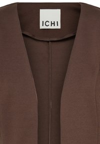 Gros plan d'un vêtement marron avec un devant ouvert et une étiquette blanche portant la mention ICHI cousue à l'intérieur de l'encolure.