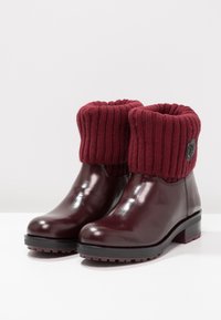 Botas de tobillo impermeables en color burdeos con un puño de punto acanalado, acabado brillante y una suela de goma negra texturizada para una mejor tracción y durabilidad.