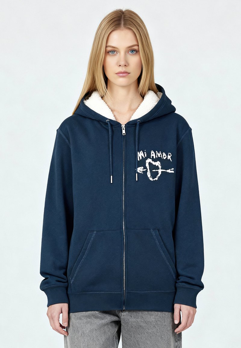 Hoodie zippé bleu marine avec une capuche en peluche doublée de blanc, comportant des poches avant et un motif blanc portant l'inscription "Mi Amor". Texture douce.