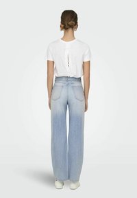 Biała bluzka z krótkim rękawem z wycięciem na plecach i detalem w postaci kokardy, zestawiona z jasnoniebieskimi spodniami typu wide-leg o wysokim stanie, noszona z białymi sneakersami.