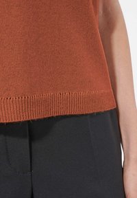 Ein rostfarbener, gestrickter Pullover, der über einer schwarzen Hose auf dem Torso und Arm einer Person sitzt, vor einem schlichten Hintergrund.