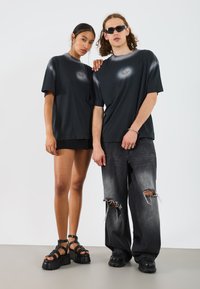 Sorte grafiske t-shirts med et centralt design, parret med slidte sorte jeans og kraftige sandaler. Glat stof med afslappet pasform.