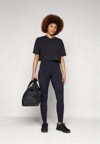 Camiseta negra recortada, leggings negros de tiro alto y zapatillas negras. La modelo sostiene una bolsa de gimnasio negra, mostrando un look atlético ajustado.