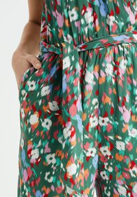 Květinový overal v živé zelené barvě, který má abstraktní barevné vzory, uvázaný pas a boční kapsy pro zvýšení funkčnosti.