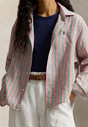 Personne portant une chemise à rayures rose et verte boutonnée sur un top côtelé bleu marine, un pantalon blanc et une ceinture tressée marron.