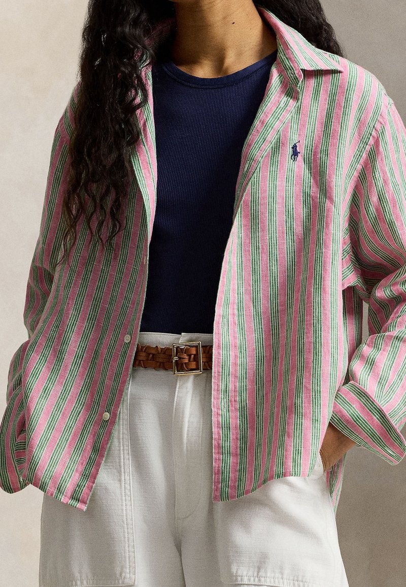 Personne portant une chemise à rayures rose et verte boutonnée sur un top côtelé bleu marine, un pantalon blanc et une ceinture tressée marron.