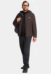 Bruine jas met rits over een zwart topje, zwarte broek en donkere schoenen. Accessoires omvatten een zonnebril en een grijze tas met een riem.