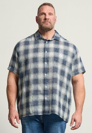 Muskelbepackter Mann, der ein kurzärmeliges, blau-weiß kariertes Hemd mit Knöpfen und blaue Jeans trägt, vor einem schlichten Hintergrund steht.