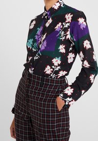 Overhemd met knopen, met een zwarte basis en een bloemmotief in wit en kleurrijke accenten. Gecombineerd met geruite broeken in rood en zwart.