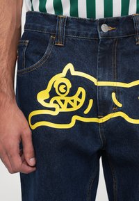 Denim jeans featuring a large yellow graphic of a playful animal design on the back pocket, with contrasting blue fabric and orange stitching.