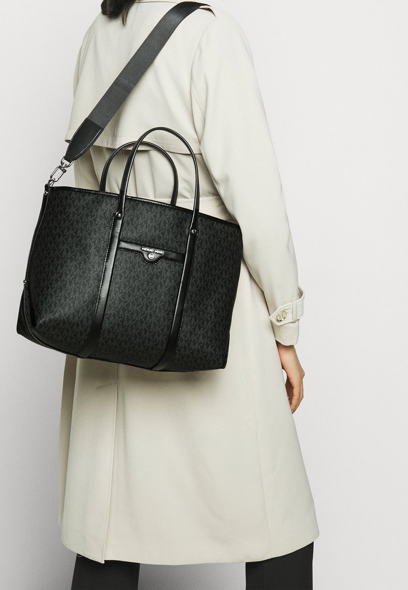 Personne portant un trench beige portant un grand sac cabas noir Michael Kors avec une bandoulière.