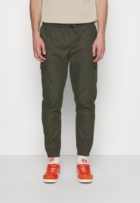 Cargo-Hosen aus dunkelgrünem Stoff, ausgestattet mit Seitentaschen, einem elastischen Bund und elastischen Bündchen an den Knöcheln, kombiniert mit orangefarbenen Sneakers.