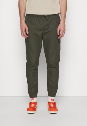 Cargo-Hosen aus dunkelgrünem Stoff, ausgestattet mit Seitentaschen, einem elastischen Bund und elastischen Bündchen an den Knöcheln, kombiniert mit orangefarbenen Sneakers.