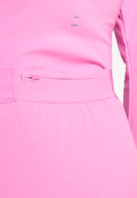 Detailní záběr jasně růžového oděvu s malou zipovou kapsou a texturovanou tkaninou se šicími detaily.