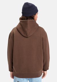 Sudadera con capucha marrón de textura suave, con un corte relajado, puños anchos y un bolsillo canguro, llevada con un gorro de punto azul marino.