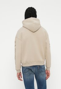 Felpa beige con cappuccio e vestibilità comoda, polsini e orlo a costine; presenta una stampa del logo lungo il braccio. Abbinata a jeans blu.