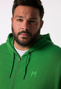 Felpa verde con zip, dotata di lacci e accessori metallici argento. Presenta un sottile logo sul petto. Texture morbida, design casual.
