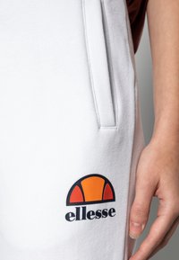 Närbild av en vit svettbyx ficka med orange och röd ellesse-logotyp, och en hand som vilar bredvid fickan.