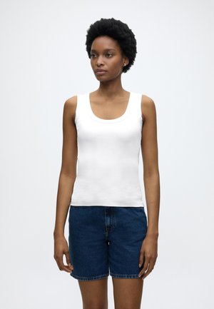 Kobieta z krótkimi naturalnymi włosami, ubrana w biały top bez rękawów i niebieskie dżinsowe szorty, stoi na tle jednolitego jasnego tła.