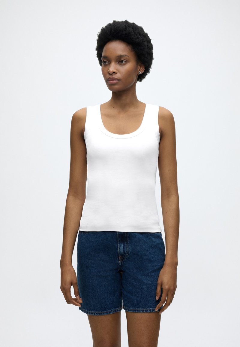 Γυναίκα με κοντά φυσικά μαλλιά, που φοράει λευκό αμάνικο τοπ και μπλε τζιν σορτς, στέκεται μπροστά σε απλό φωτεινό φόντο.