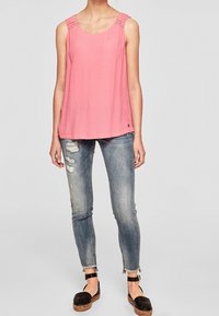 Top sin mangas de color rosa con detalles de hombros plisados, combinado con jeans skinny azules desgastados y sandalias de plataforma negras con acentos trenzados.