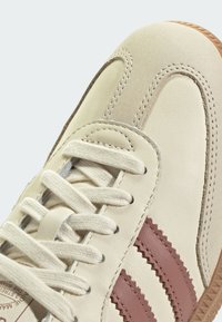 Kremhvit skinnsneakers med tre burgunderrøde taggete striper, lyse beige semskede aksenter, flate lisser og en gummisåle.