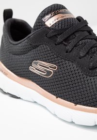 Svart mesh-sneaker med roséguld-accenter, som har en lättviktsdesign, texturerat överdel och en vit gummisula med ett zigzag-mönster.