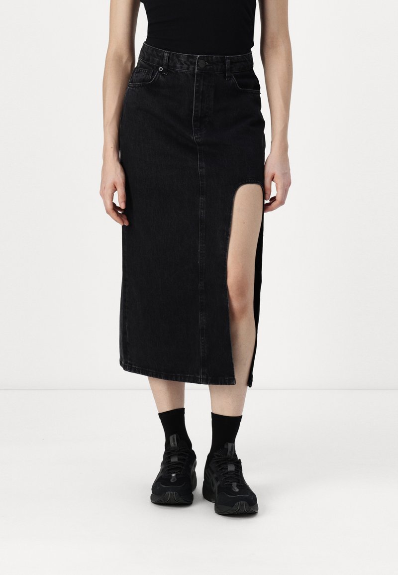 Saia de denim preta com uma fenda até a coxa, apresentando um fecho de botão na frente e bolsos. Usada com meias pretas e ténis.